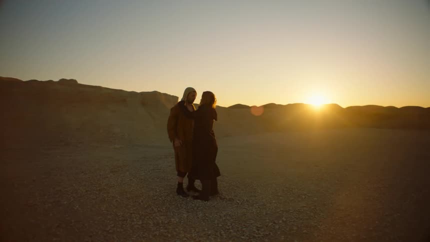 A man and woman stand in cloaks in the desert