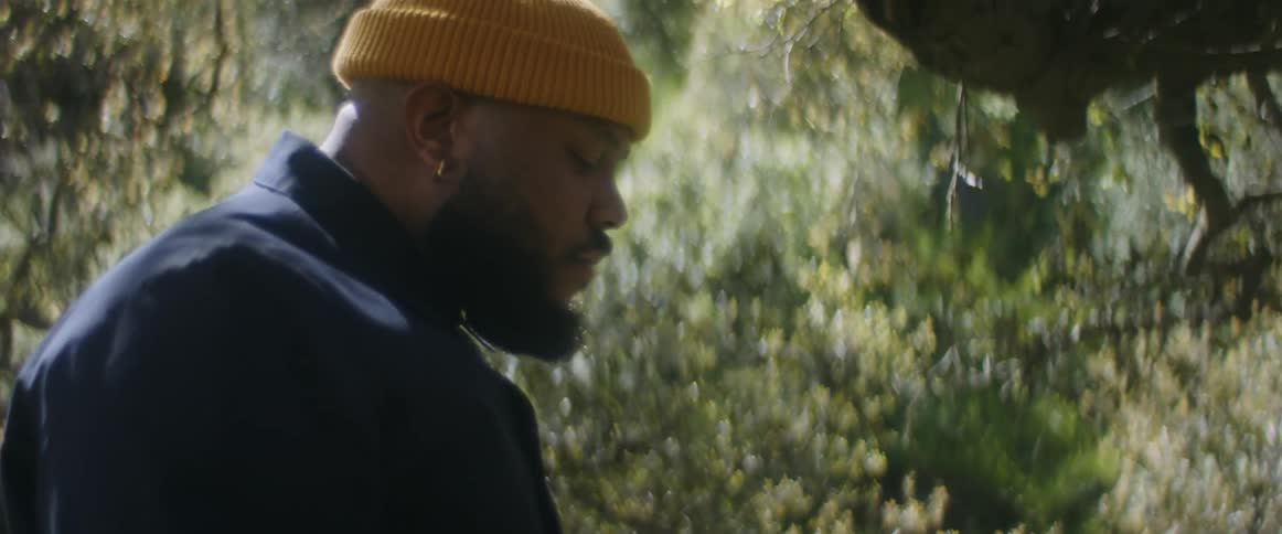 A man is wearing a coat and a yellow knit cap as he walks near trees in a field on a sunny day