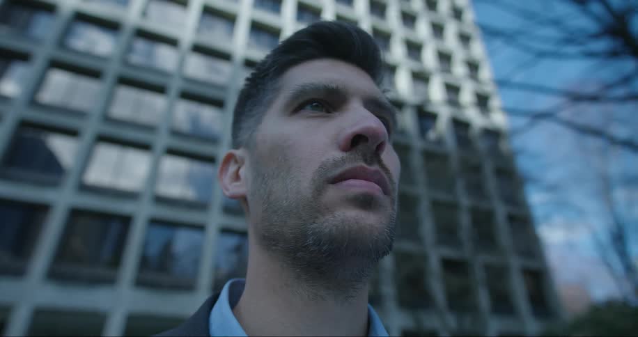 A close up of a mans face staring out in front of him while he stands in front of a tall building