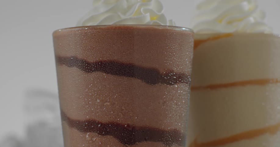 Two blended coffee drinks with whipped cream topping one is mocha the other is caramel on a table surrounded by coffee beans