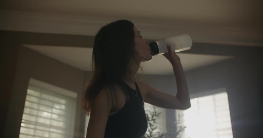 A woman stands and drinks from a water bottle and reaches her arm out to set it back down