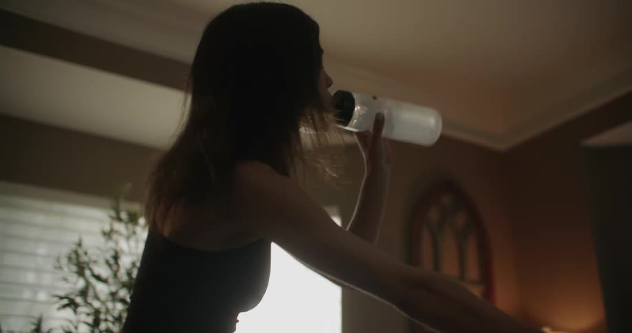 A woman drinking from a water bottle while exercising on a stationary bike at home