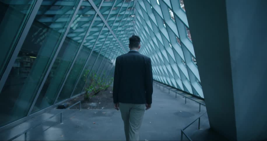 The backside of a man as he walks down a corridor in a building with his head down looking at the ground