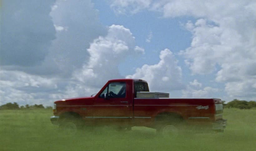 A red pickup truck driving through a grassy field near a tree
