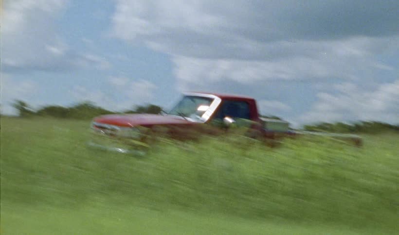 Red truck drives through field with trees on the horizon under a cloudy sky