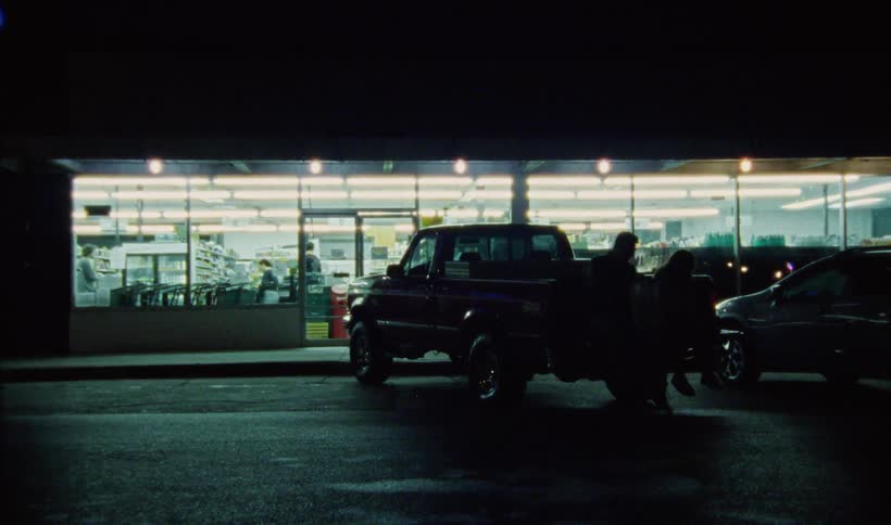Man comes out of a supermarket into the parking lot at nighttime where he joins a young woman on the tailgate of his truck