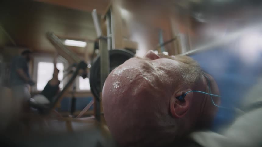 A man at a gym bench presses heavy weights while laying on a bench and listening to earbuds