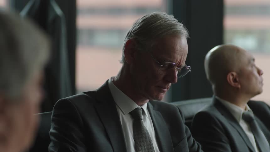 Two businessmen sitting next to each other in suits one writing and looking up every so often