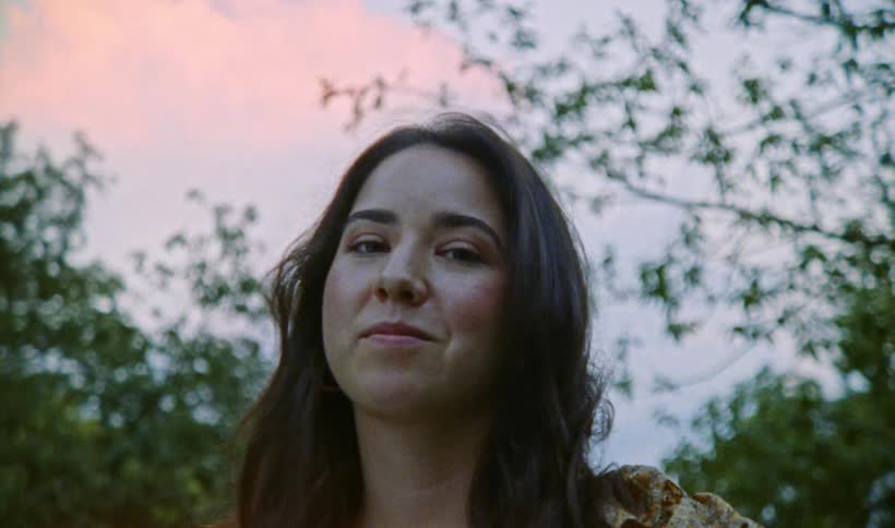 Woman with floral dress looks down at camera near the trees at dusk