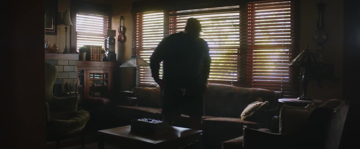 A man stands up from the couch under a window with blinds