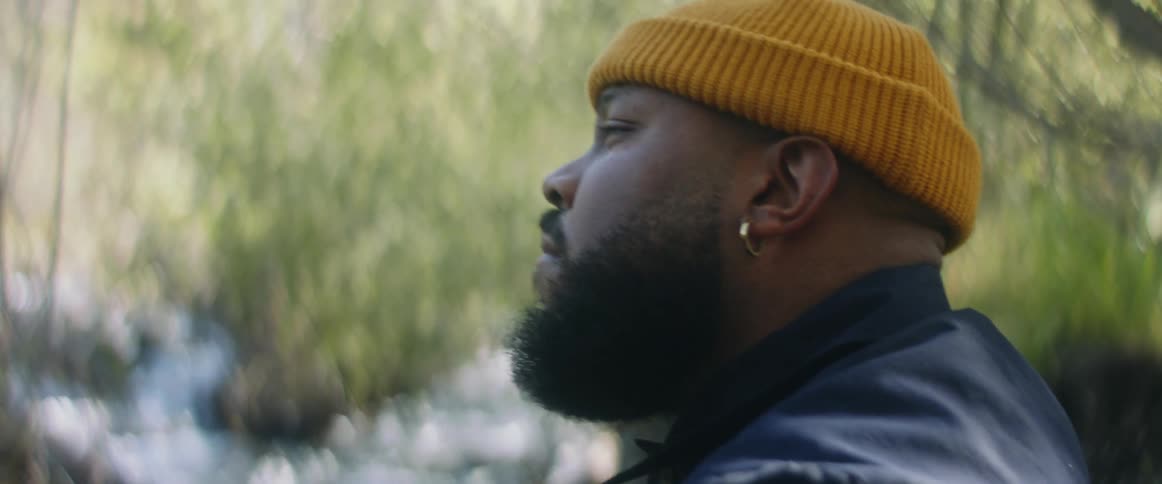 Man with yellow beanie earrings beard and blue jacket stands by stream in the forest