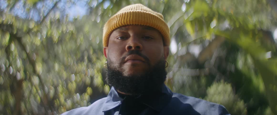 A man with a beard and yellow beanie blinks as he stands in front of trees