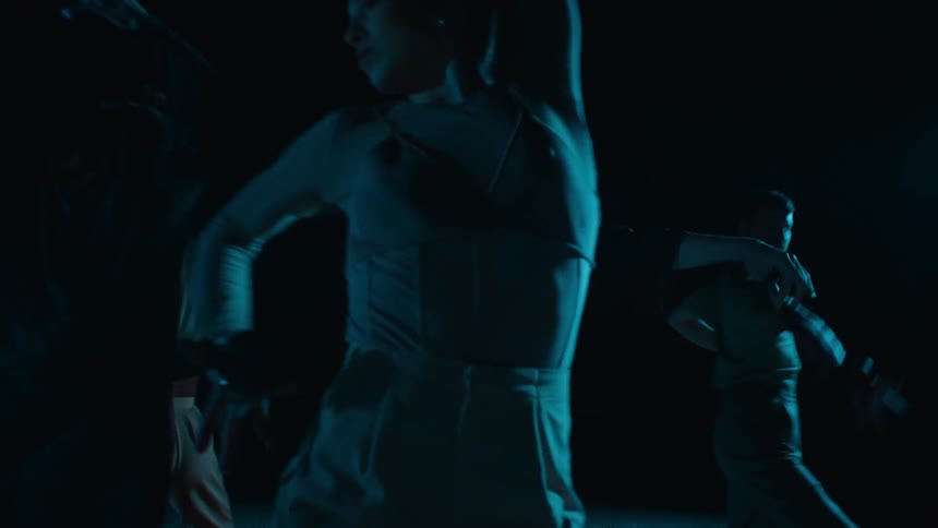 A man dances vigorously with a group of dancing women around him dimly lit by blue light