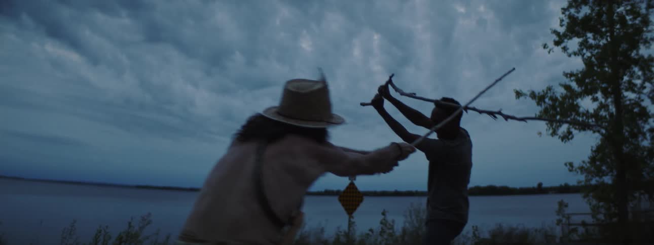 The silhouette of two men play fighting with sticks outside near a lake at night