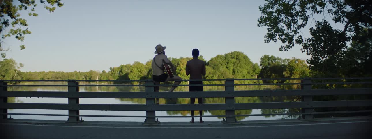 Two men are on a bridge over a lake making music on a sunny day
