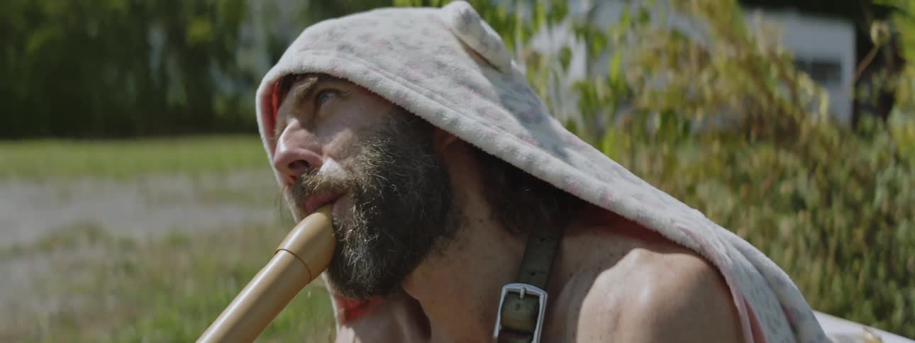 A man with a hooded jacket on his head while he plays a wood instrument looking ahead of him without a shirt on
