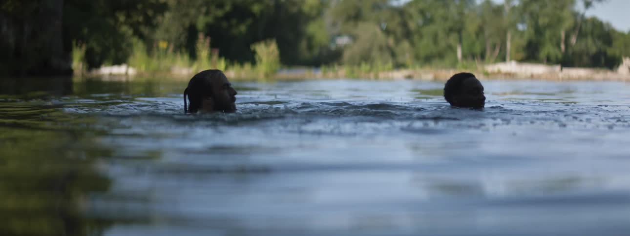 Two men are playfully swimming in a lake in a forested area during the daytime