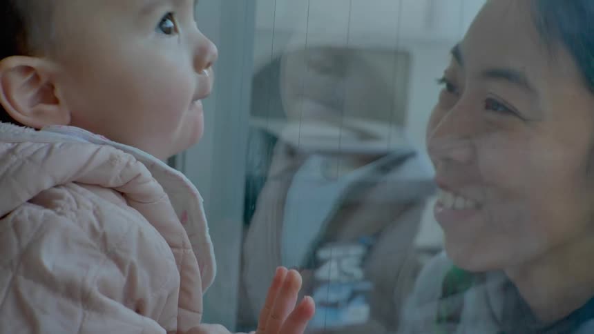 A close up of a womans face as sh makes kissing faces at the baby on the other side of the window