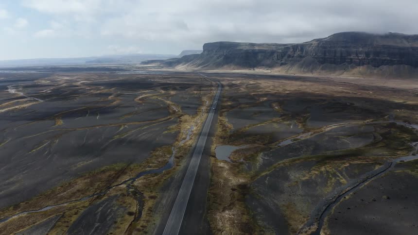 A plateau of badlands with a single road traveling through it from a drone view with mountains in the distance