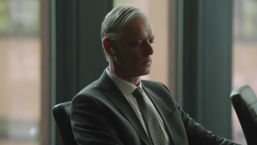 An old man sits worried at a board meeting listening to something on his earbuds and ignoring the people talking next to him