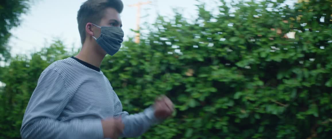 Man with a long sleeve shirt wears a grey face mask and walks to a building in a garden