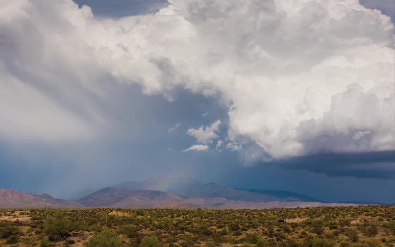 White clouds and a rainbow are seen in time lapse as they move over some hills