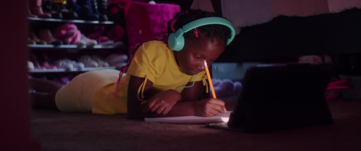 Young female wearing headphones lies on her stomach on the floor of a bedroom illuminated by the light of a laptop while using a pencil to write in a notebook