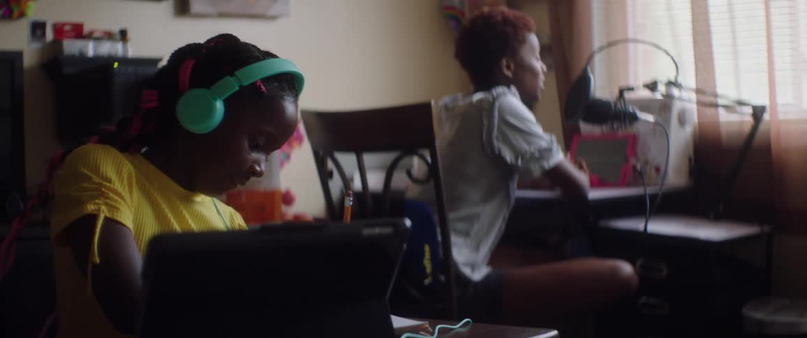 A young girl sits in front of her laptop while her mom sits at another desk and talks in to a microphone