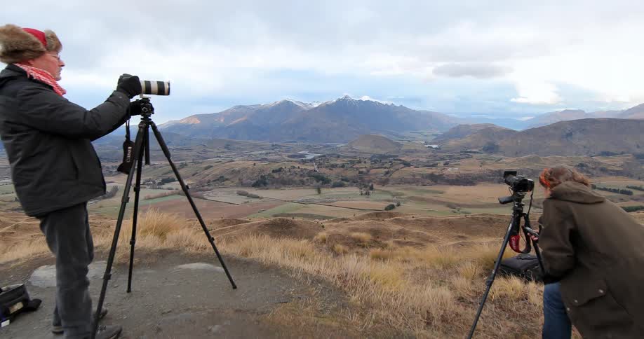 Two people have tripods set up on a hill overseeing a large hilly area below