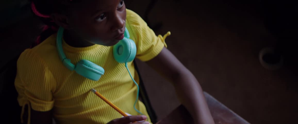 Black child with a yellow t shirt and turquoise headphones sitting at the desk and writes in a notebook
