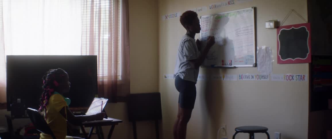 Female student sits at a desk while a teacher writes on a whiteboard
