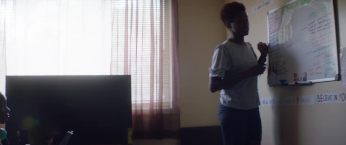Female teacher writes on a white board as a young black girl sits at a desk
