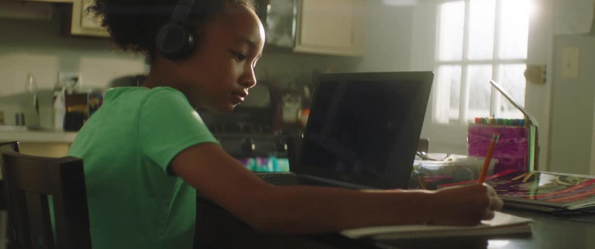 Black girl with a green shirt and headphones sitting at the table looks at a laptop and writes down in a notebook in the kitchen