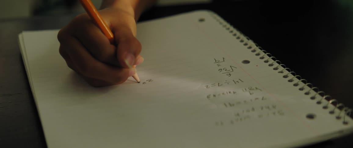 A person sitting by a desk writing on a notebook with a pencil