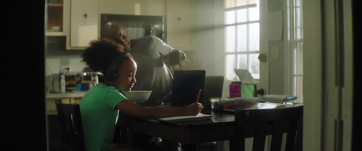 Black teenager sits at a kitchen table with a laptop as her father sits down with a cup of coffee
