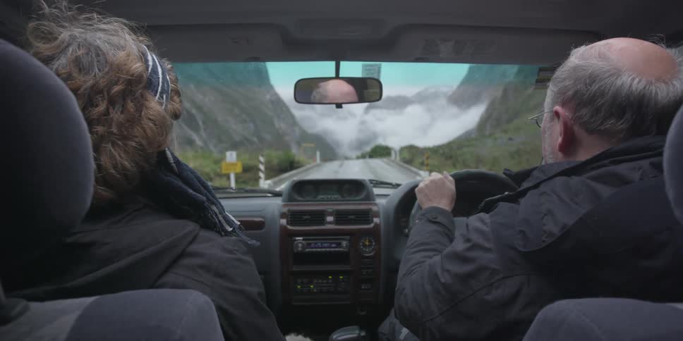 A man and woman drive down a road by a mountain