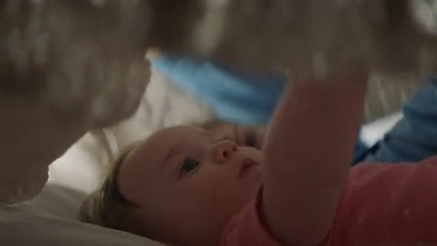 A baby is lying on a bed as his mother lying next to him pretends to completely cover them both with a blanket