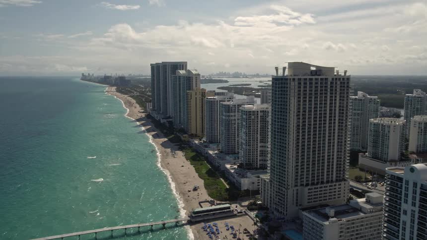 Approaching a pier aerial footage captures the passing beachfront high-rises and stunning coastline of Sunny Isles Beach in Florida