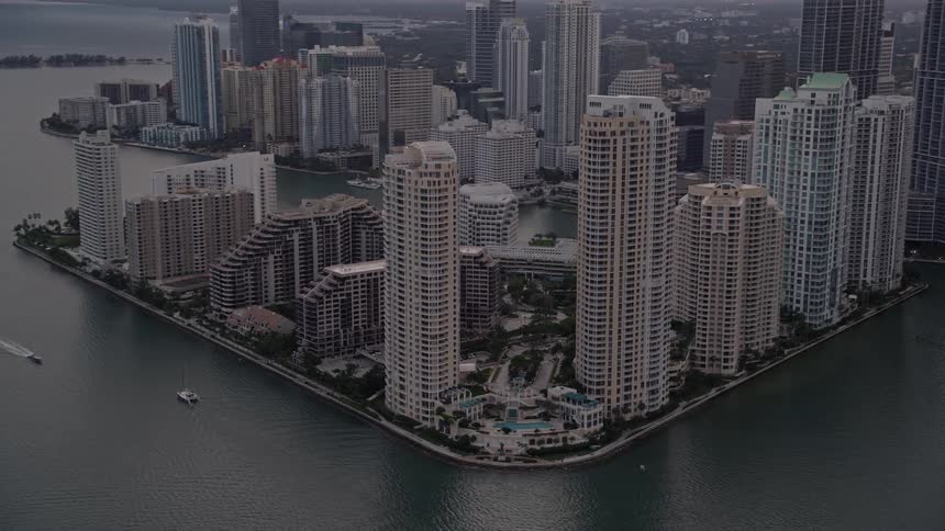 Aerial footage of high rise buildings on the brick-lined coast of downtown Miami during sundown with city skyline in the background and boats passing through