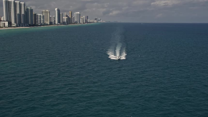An aerial view captures a motorboat racing across the ocean near Sunny Isles Beach with partly cloudy skies and coastal city in the background