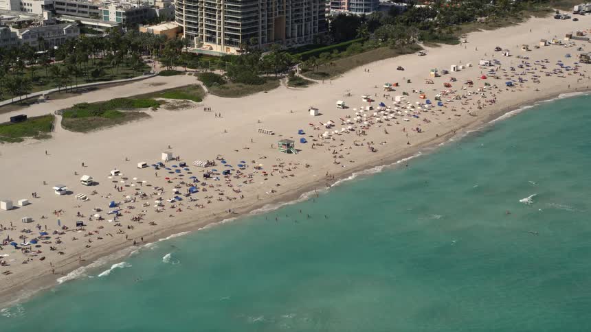 Aerial footage captures a lively beach scene in South Beach Florida with crowds of sunbathers and beachgoers enjoying various recreational activities by the shoreline