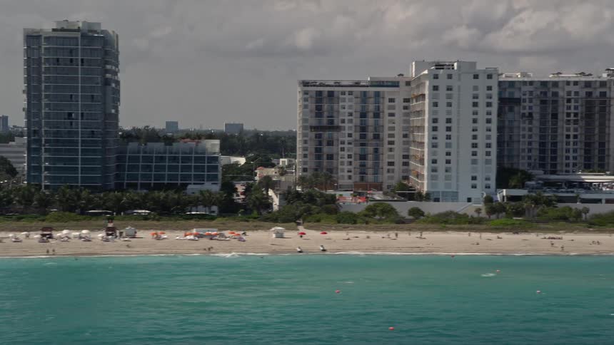 Aerial footage of a row of beachfront condominiums in Miami Beach Florida with a crowd of people enjoying the shoreline below
