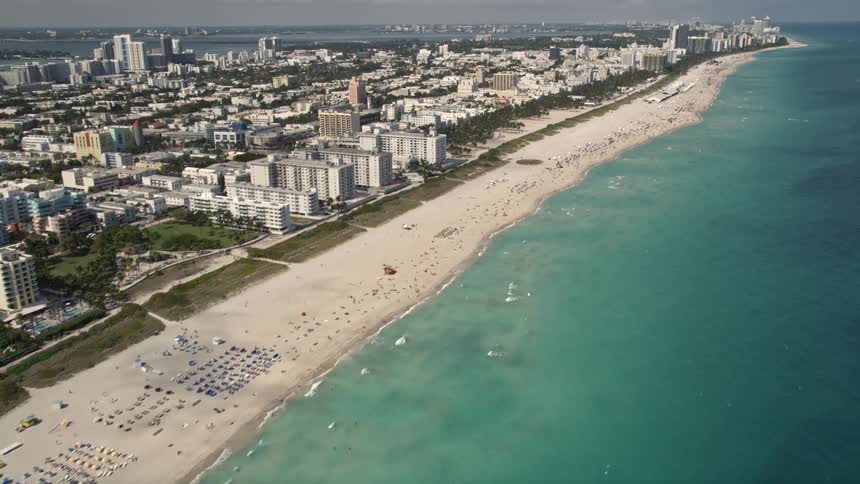 Aerial footage tilting up from sunbathers on South Beach to show the partly cloudy skyline of Miami and the residential buildings behind it