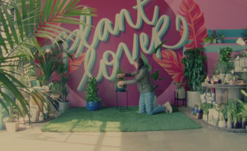 A woman knelt down on turf fixing a plants leaves that is on a stand posed for a picture