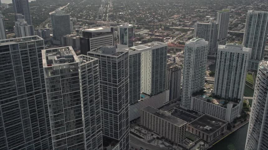 A drone approaches high-rise complex in Downtown Miami as it hovers over the Miami River and captures the modern buildings and city skyline in the background against a cloudy sky