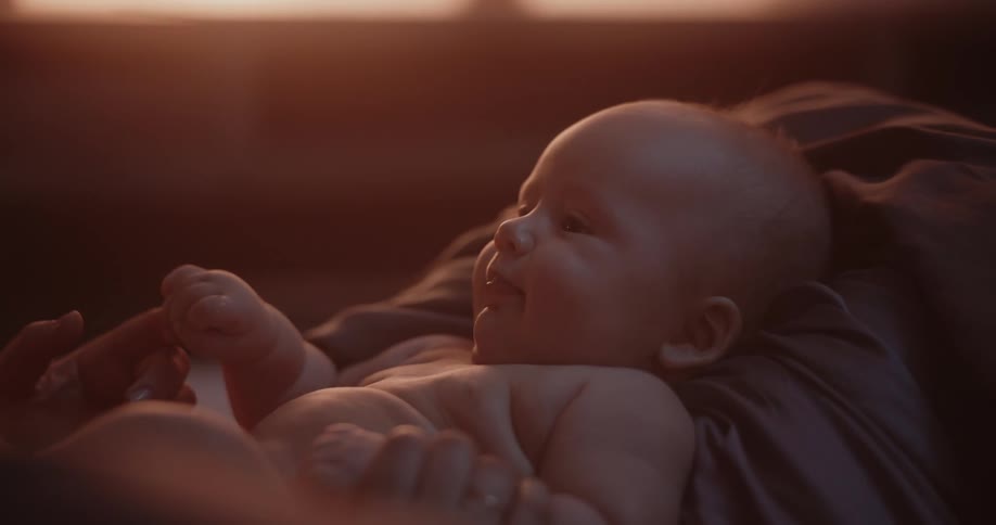 A baby lying against a pillow looks at someone and grabs their finger