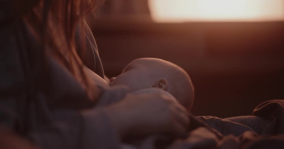 Mother with brown hair breastfeeds baby while the sun streams through windows in room
