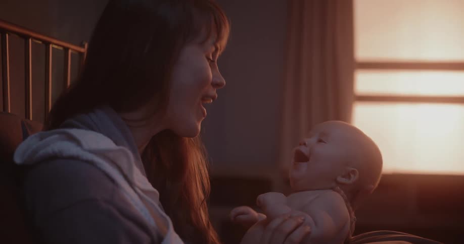 Mother sitting on bed plays with her baby as red light shines through nearby window