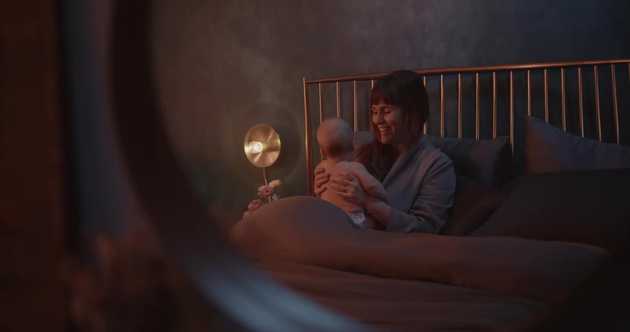 Mother with bangs sits on bed holding newborn baby with diaper in bedroom