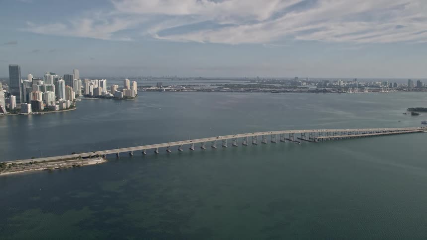 Aerial view of traffic crossing a long bridge across the water from downtown skyscrapers revealing the shoreline and Downtown Miami in Florida on a sunny day